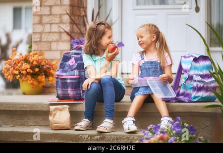 Élèves de l'école primaire avec des boîtes à lunch entre les mains. Les filles avec des sacs à dos mangent des fruits près du bâtiment à l'extérieur. Début des leçons. Premier jour de f Banque D'Images