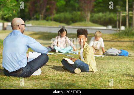 Portrait complet d'un garçon afro-américain souriant, assis sur une pelouse verte et regardant l'appareil photo tout en profitant de la classe en plein air sous la lumière du soleil, dans un espace de copie Banque D'Images