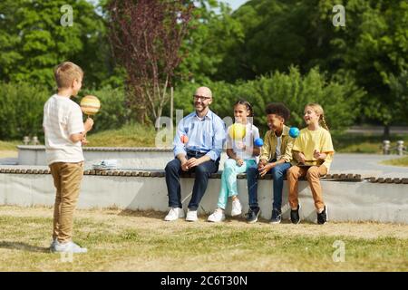 Portrait complet d'un professeur souriant écoutant un garçon en donnant une présentation sur l'astronomie tout en étant assis avec un groupe d'enfants et en appréciant l'outdo Banque D'Images