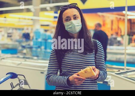 une femme dans un masque médical de protection dans un supermarché choisit le fromage. pandémie et covid-19 concept. Main de l'acheteur avec un morceau de fromage dans le magasin Banque D'Images