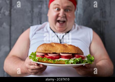 un jeune homme de caucase en surpoids empiète sur un grand sandwich à haute teneur en calories, l'homme affamé veut manger, il ne s'occupe pas de gain de poids Banque D'Images