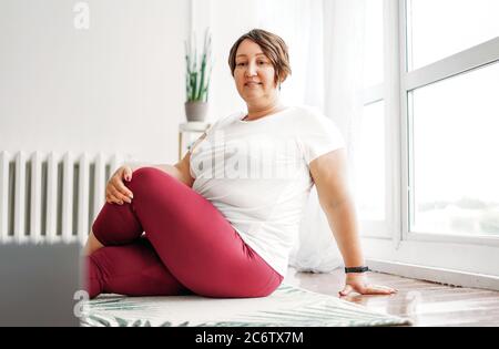 Adulte charmante femme brune plus taille corps pratique positive yoga avec ordinateur portable à la maison Banque D'Images