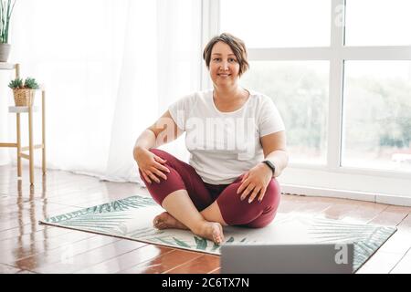 Adulte charmante femme brune plus taille corps pratique positive yoga avec ordinateur portable à la maison Banque D'Images