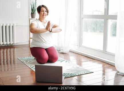 Adulte charmante femme brune plus taille corps pratique positive yoga avec ordinateur portable à la maison Banque D'Images