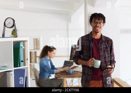 Portrait à la taille haute d'un homme afro-américain souriant tenant une tasse et regardant l'appareil photo tout en se tenant contre le mur dans un intérieur de bureau contemporain, copie Banque D'Images