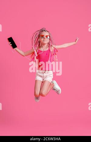 Belle petite fille avec des dreadlocks sautant avec le téléphone mobile dans la main et écouter de la musique sur casque sur fond rose. Banque D'Images