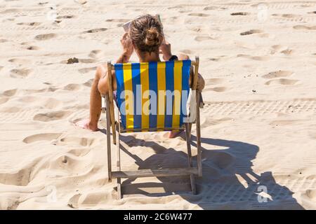 Homme assis dans un transat lisant un livre sur la plage à Bournemouth, Dorset UK, par une chaude journée ensoleillée en juillet - vue arrière Banque D'Images