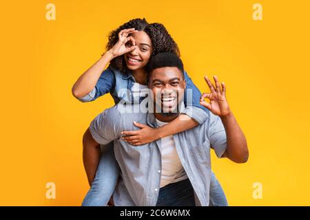 Ambiance ludique. Couple noir montrant des signes OK pendant que Guy piggydorarrière petite amie Banque D'Images