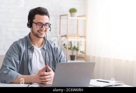 Apprendre l'anglais en ligne. Homme souriant avec un casque regardant un ordinateur portable, parlant avec un professeur Banque D'Images