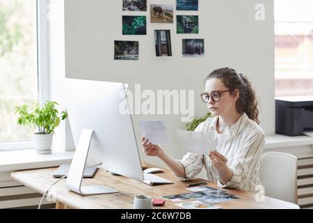 Portrait de jeune femme moderne tenant des photographies pour la revue en travaillant à PC dans le bureau blanc, espace de copie Banque D'Images