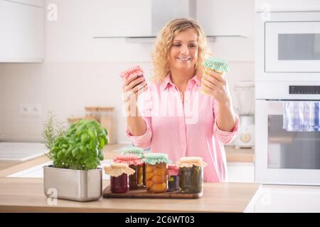 Femme regardant des conserves maison fraîches dans ses mains avec plus de cornichons dans des pots en face d'elle. Banque D'Images
