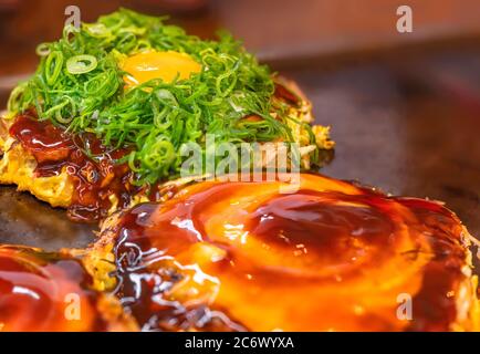 Gros plan sur un œuf frit recouvert de gros oignons frits hachés sur une omelette japonaise okonomiyaki cuite sur une plaque chaude et brossée en cercle Banque D'Images