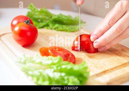 Les mains élégantes d'une jeune fille coupent la tomate rouge juteuse en deux sur une planche à découper en bois. Préparation des ingrédients et des légumes. Produits biologiques Banque D'Images