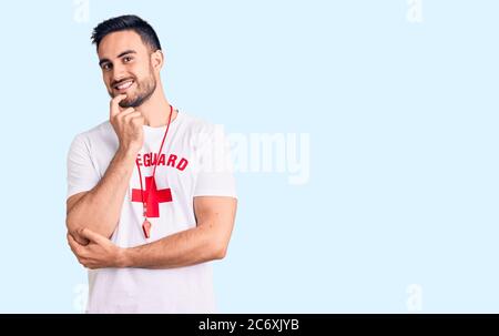 Jeune homme beau portant un uniforme de maître-nageur en regardant avec confiance la caméra avec le sourire avec les bras croisés et la main levée sur le menton. En pensant positif. Banque D'Images