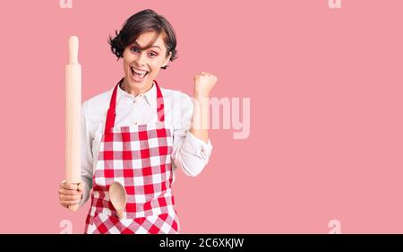 Belle jeune femme aux cheveux courts portant un tablier boulanger professionnel tenant le rouleau de pétrissage criant fier, célébrant la victoire et le succès très exc Banque D'Images