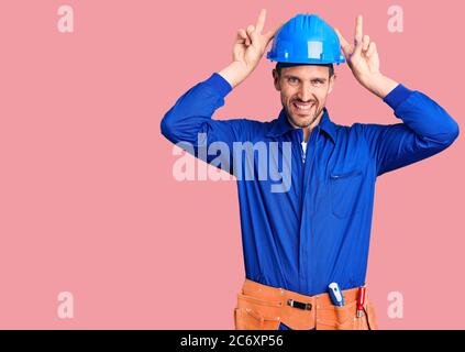 Jeune homme beau portant l'uniforme de travailleur et le casque de sécurité posant drôle et fou avec les doigts sur la tête comme des oreilles de lapin, souriant gai Banque D'Images