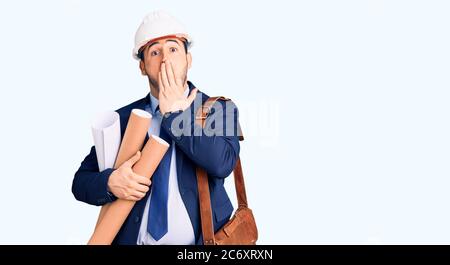 Jeune homme hispanique portant un casque d'architecte et un sac en cuir tenant des bleus couvrant la bouche avec la main, choqué et peur pour erreur. Surpris ex Banque D'Images