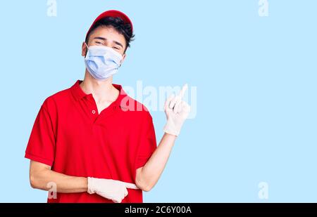 Jeune homme hispanique portant l'uniforme de livraison et le masque médical souriant heureux pointant avec la main et le doigt sur le côté Banque D'Images