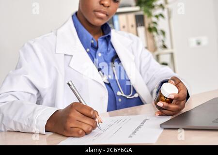 African american médecin écrivant prescription tenant flacon de pilules s'asseoir à un bureau. Banque D'Images
