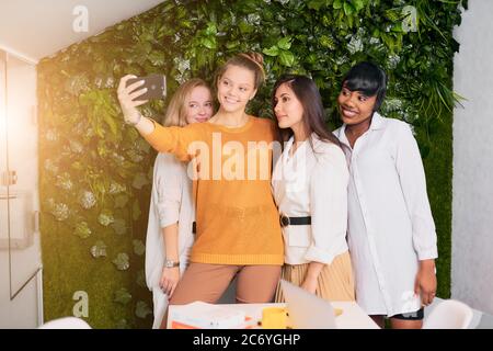 Prenons un selfie, une photo sur le lieu de travail. Les jeunes femmes caucasiennes et africaines sont heureuses ensemble en pause au bureau. Loisirs Banque D'Images