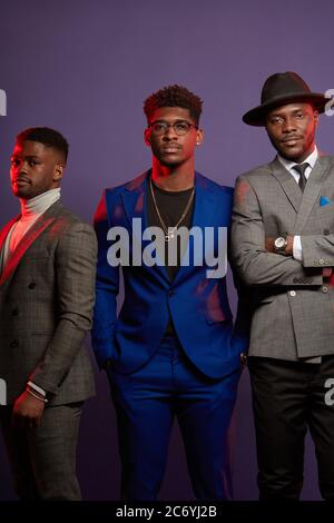 Groupe de trois amis africains vêtus de costumes élégants pose isolés en studio sur fond violet foncé. Un des gars porte un chapeau. Classe Banque D'Images