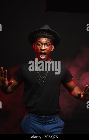 Un jeune africain portant un chapeau élégant et un t-shirt noir regardant un appareil photo avec une large bouche ouverte criant sur un fond sombre fumé garde les mains comme si h. Banque D'Images
