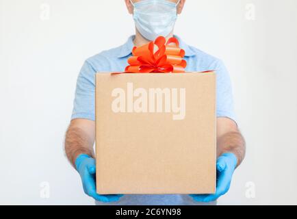 Un homme en gants médicaux et un masque tient une boîte avec un ruban rouge et un noeud sur un fond blanc. Vente. Cadeau. Coronavirus, quarantaine, vente Banque D'Images