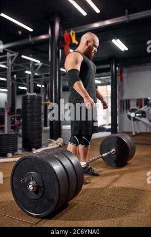 Vue rapprochée de barbell en métal lourd, couché sur un sol marron dans la salle de gym, homme chauve avec une barbe épaisse qui regarde loin, se préparant à l'entraînement, culturiste de sport c Banque D'Images