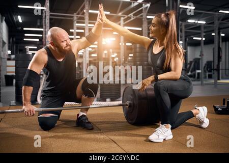 Jeune couple actif faisant le pari dans la salle de gym lumineuse, levant les mains, touchant les paumes dans les airs, prêt à commencer l'entraînement dans l'électronique moderne de fitness Banque D'Images