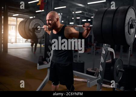 Powerlifter chaudrée sans cheveux avec une barbe épaisse vêtue de vêtements de sport noirs, essayant de lever la barbell lourde, faisant l'effort, l'entraînement dur, la préparation Banque D'Images