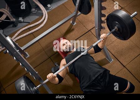 Beau chauve avec une barbe brune épaisse allongé sur un banc avec une barbell lourde dans les mains, poids de levage dans la salle de gym, apprécier la musculation, faire des efforts, Banque D'Images