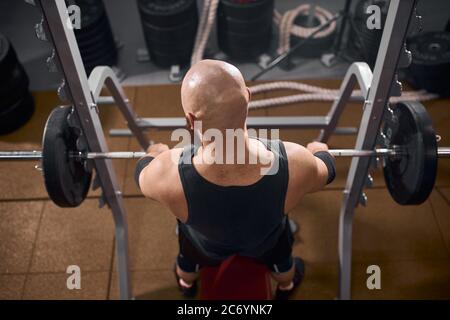 Vue arrière d'un homme chauve sans cheveux assis devant une barbell en fer lourd, en gardant les mains sur la barre métallique, en préparant la presse sur banc, vue à angle élevé, intérieur s. Banque D'Images