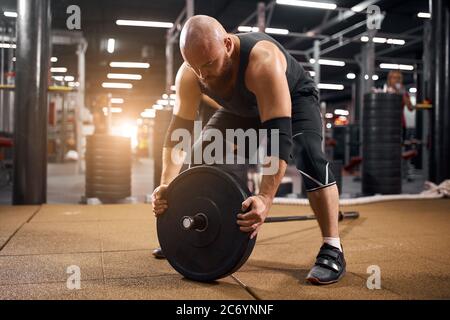 Culturiste sportif dans une salle de gym lumineuse avec une plaque de musculation entre les mains, préparant la barbell pour l'entraînement, regardant vers le bas, exprimant dur, fit p Banque D'Images