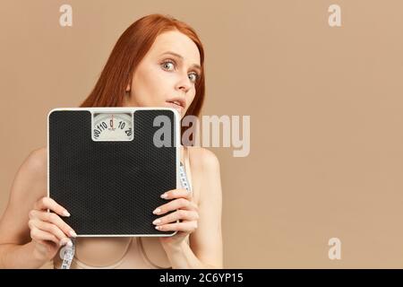 Timide effrayé rouillé femme skinny à cheveux rouges avec les yeux bougés, se cachant face derrière les balances mécaniques, isolé sur fond beige Banque D'Images