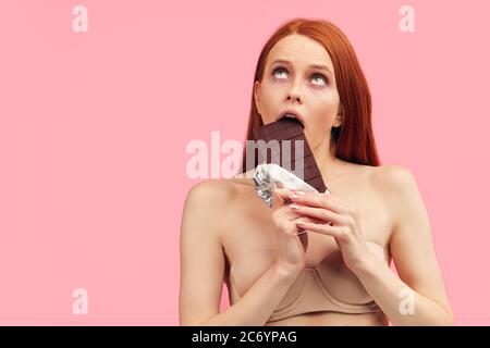 Bonne jeune femme maigre épuisé par des régimes sévères, tenant la barre de chocolat, posant isolé sur rose. L'échec de régime de la femme rouge emaciée est en train de manger Banque D'Images