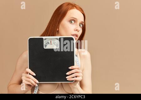 Timide effrayé rouillé femme skinny à cheveux rouges avec les yeux bougés, se cachant face derrière les balances mécaniques, isolé sur fond beige Banque D'Images