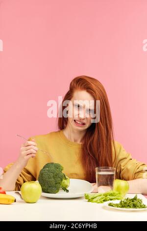 Triste rouge-cheveux femme se bouclent les lèvres tandis que regarde les légumes pour le déjeuner, fatigué de régime végétarien strict et hate la verdure. Une adolescente tient le brocco Banque D'Images