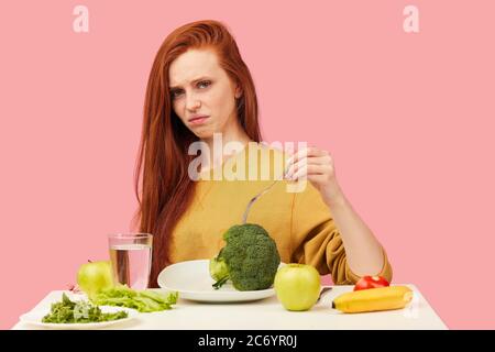 Triste femme aux cheveux rouges dans le mauvais temps gardant un régime végétarien strict fatiguée des restrictions et déteste la verdure. Une adolescente tient le brocoli sur la fourchette Banque D'Images