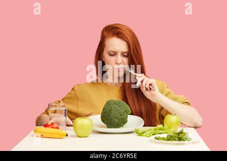 Femme aux cheveux rouges dans le mauvais temps gardant un régime végétarien strict fatiguée des restrictions et déteste la verdure. Une adolescente tient le brocoli sur la fourchette Banque D'Images