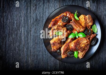 Côtes de porc braisées aux pruneaux servies sur une assiette noire, feuilles de basilic sur fond de bois foncé, vue du dessus, espace de copie Banque D'Images