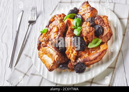 Petites côtes de porc braisées avec pruneaux, bouillon, oignon et ail, décorées de basilic frais, servies sur une assiette blanche sur un backgrou en bois blanc Banque D'Images