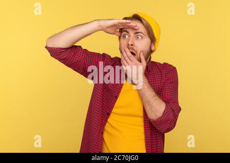 Vision lointaine. Un gars hipster stupéfié en bonnet beanie et une chemise à carreaux tenant la main sur les yeux regardant sur la longue distance avec surprise d'expression choquée, Banque D'Images