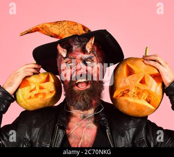 Halloween – concept de fête effrayante. Monstre avec décoration d'octobre. Démon avec des cornes et le visage suspect tient Jack o lanternes. Homme portant un maquillage effrayant et un chapeau de sorcières tient des citrouilles sur fond rose Banque D'Images