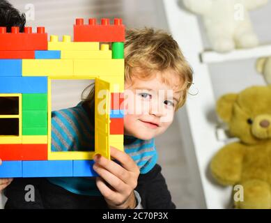 Un garçon joue avec des jouets sur fond de salle de bébé. Concept de jeux d'enfance et actifs. Cache-enfants derrière le mur du bâtiment en blocs de plastique. Enfant avec visage heureux tient coloré jouet briques construction Banque D'Images