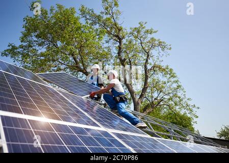 Deux travailleurs dans un casque de protection sur une plate-forme en acier installant un panneau solaire solaire photovoltaïque lourd sur fond vert et bleu ciel. Installation du système de panneau solaire extérieur, concept de travail dangereux. Banque D'Images