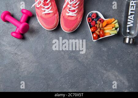 Fitness concept, des sneakers roses, des haltères, une bouteille d'eau et une assiette en forme de coeur avec des légumes et des baies sur un fond de beton, vue du dessus, sain Banque D'Images