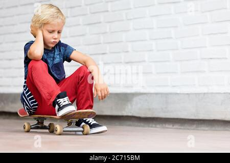 Le patineur qui a l'air malheureux et contrarié s'assoit sur le skateboard avant que les enfants s'entraîne dans le parc de skate. Vie familiale active, activités de loisirs en plein air Banque D'Images