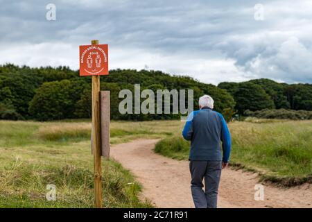 East Lothian, Écosse, Royaume-Uni, 13 juillet 2020. Météo au Royaume-Uni : couvert à la plage de Yellowcraig. Des mesures de distanciation sociale sont en place sur le trajet entre le parc automobile et la plage, tandis qu'un homme senior marche le long du sentier pendant la pandémie du coronavirus Covid-19 avec un avis de rester à 2 mètres d'écart Banque D'Images