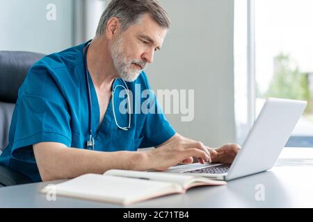 Un médecin mature travaillant sur un ordinateur portable, assis dans un bureau médical. Banque D'Images