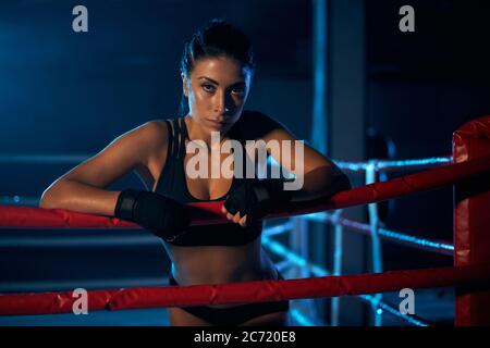 kickboxer femme professionnelle sérieuse jeune portant des vêtements de sport et des bandages noirs sur les mains, posant dans une atmosphère sombre. Chasseur ayant le repos après l'entraînement, se pencher sur la corde et regarder la caméra. Banque D'Images